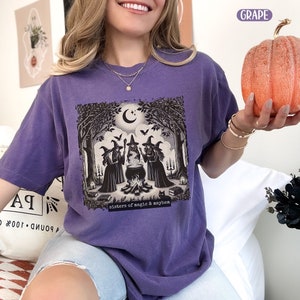 Puede incluir: Camiseta morada con un gr&aacute;fico de tres brujas de pie alrededor de un caldero en un bosque. El texto "Sisters of magic & mayhem" est&aacute; impreso debajo del gr&aacute;fico.