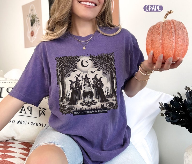 Puede incluir: Camiseta morada con un gr&aacute;fico de tres brujas de pie alrededor de un caldero en un bosque. El texto "Sisters of magic & mayhem" est&aacute; impreso debajo del gr&aacute;fico.