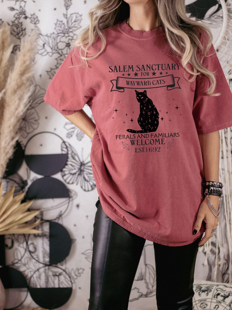 Może przedstawiać: Oversizowy t-shirt w kolorze bordowym z czarnym nadrukiem graficznym, na kt&oacute;rym widnieje napis "Salem Sanctuary for Wayward Cats" z czarną sylwetką kota i gwiazdami. Tekst pod kotem brzmi "Ferals and Familiars Welcome Est 1692".