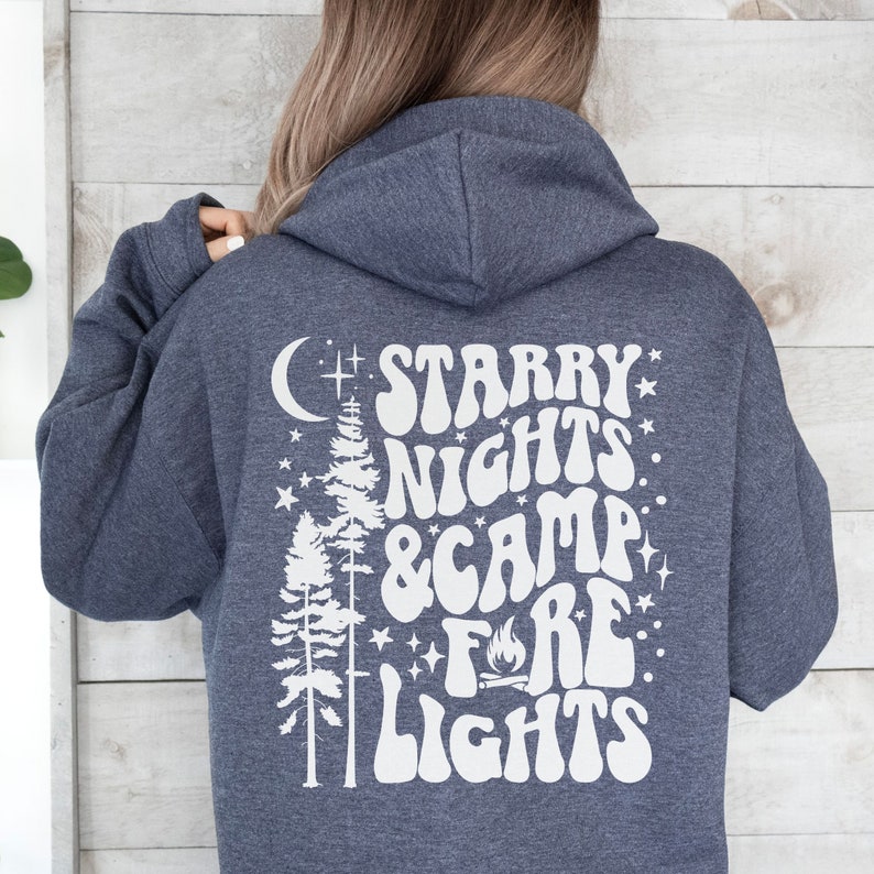 May include: A dark blue hooded sweatshirt with a white graphic design on the back. The design features a crescent moon, two pine trees, stars, and the text "Starry Nights & Camp Fire Lights" in a retro font.