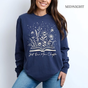 May include: Midnight blue long-sleeve sweatshirt with a floral and celestial design. The graphic features an open book with flowers and stars, and the text "Just One More Chapter".