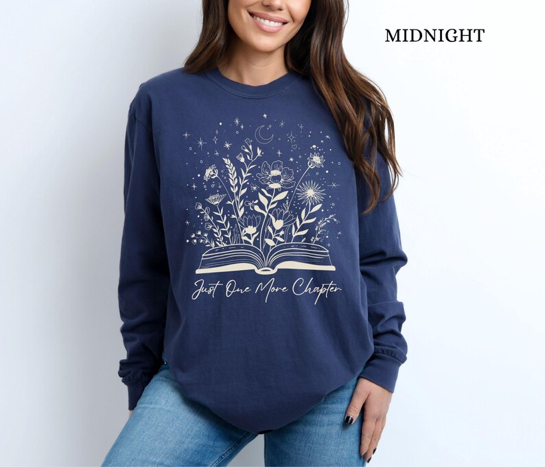May include: Midnight blue long-sleeve sweatshirt with a floral and celestial design. The graphic features an open book with flowers and stars, and the text "Just One More Chapter".