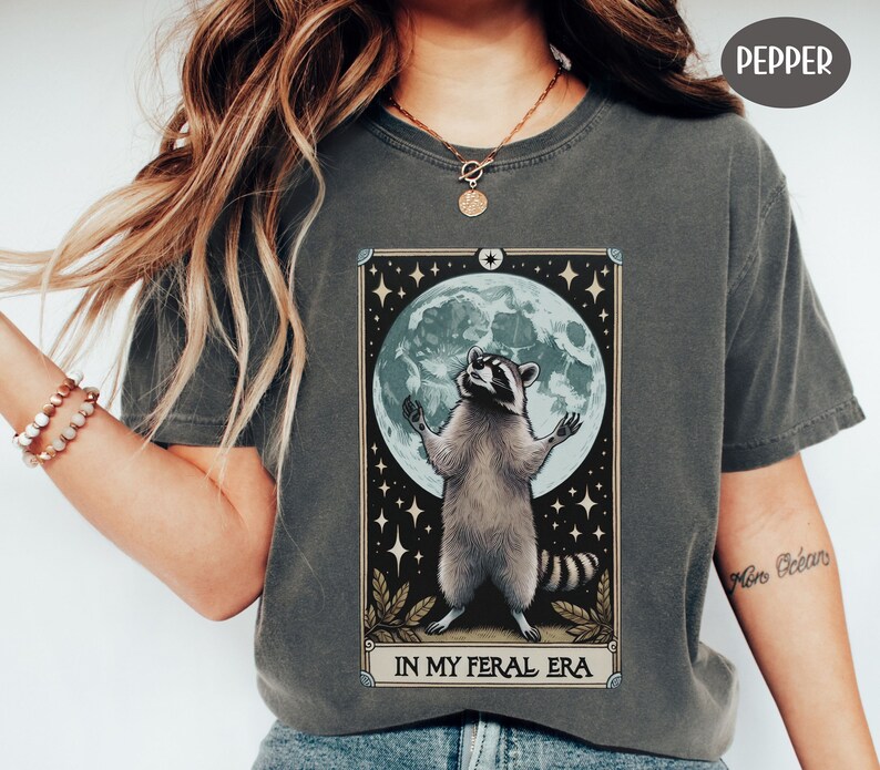 May include: A dark gray t-shirt with a graphic of a raccoon standing in front of a full moon with stars. The text "In My Feral Era" is printed below the graphic.