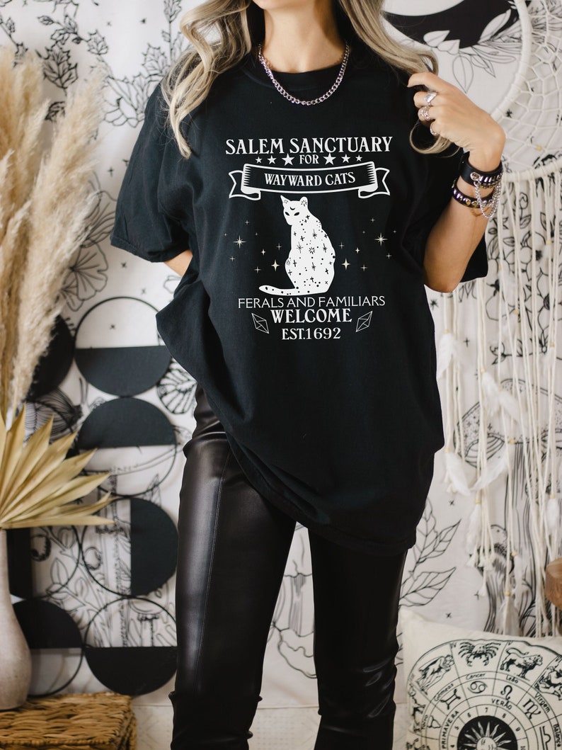 Może przedstawiać: Czarny T-shirt z białym nadrukiem przedstawiającym kota z gwiazdami i tekstem "Salem Sanctuary for Wayward Cats, Feral and Familiars Welcome, Est. 1692."