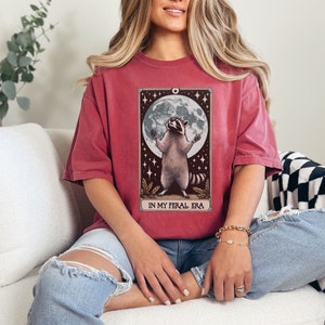 May include: A red t-shirt with a graphic of a raccoon standing on its hind legs with its paws raised in the air. The raccoon is in front of a full moon and stars. The text "IN MY FERAL ERA" is printed below the raccoon.