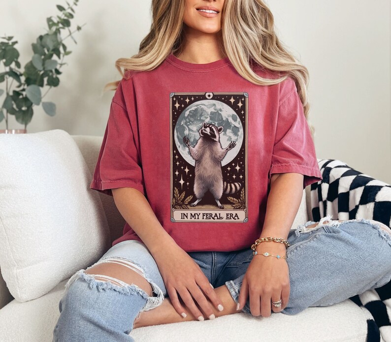 May include: A red t-shirt with a graphic of a raccoon standing on its hind legs with its paws raised in the air. The raccoon is in front of a full moon and stars. The text "IN MY FERAL ERA" is printed below the raccoon.