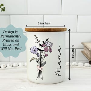 May include: A white ceramic jar with a bamboo lid, 3 inches in diameter and 4 inches tall. It features a floral design with purple and pink flowers and the word "Mama" in black script. The design is permanently printed on the glass.