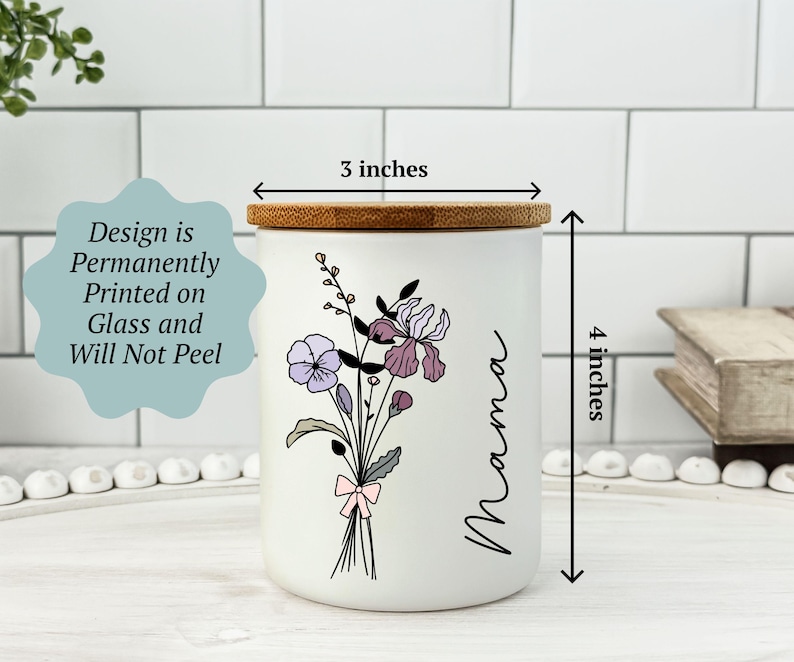 May include: A white ceramic jar with a bamboo lid, 3 inches in diameter and 4 inches tall. It features a floral design with purple and pink flowers and the word "Mama" in black script. The design is permanently printed on the glass.