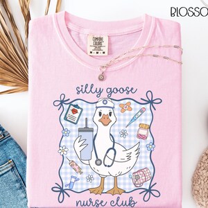 May include: A light pink t-shirt with a graphic of a goose wearing a nurse's hat and stethoscope, holding a drink. The text "silly goose nurse club" is displayed. The shirt has a Comfort Colors tag and a gold necklace.