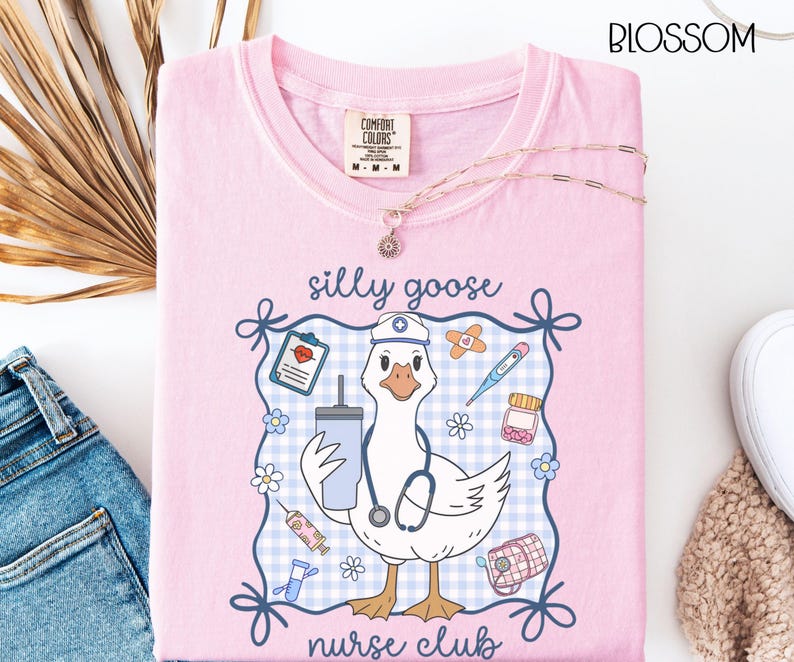 May include: A light pink t-shirt with a graphic of a goose wearing a nurse's hat and stethoscope, holding a drink. The text "silly goose nurse club" is displayed. The shirt has a Comfort Colors tag and a gold necklace.