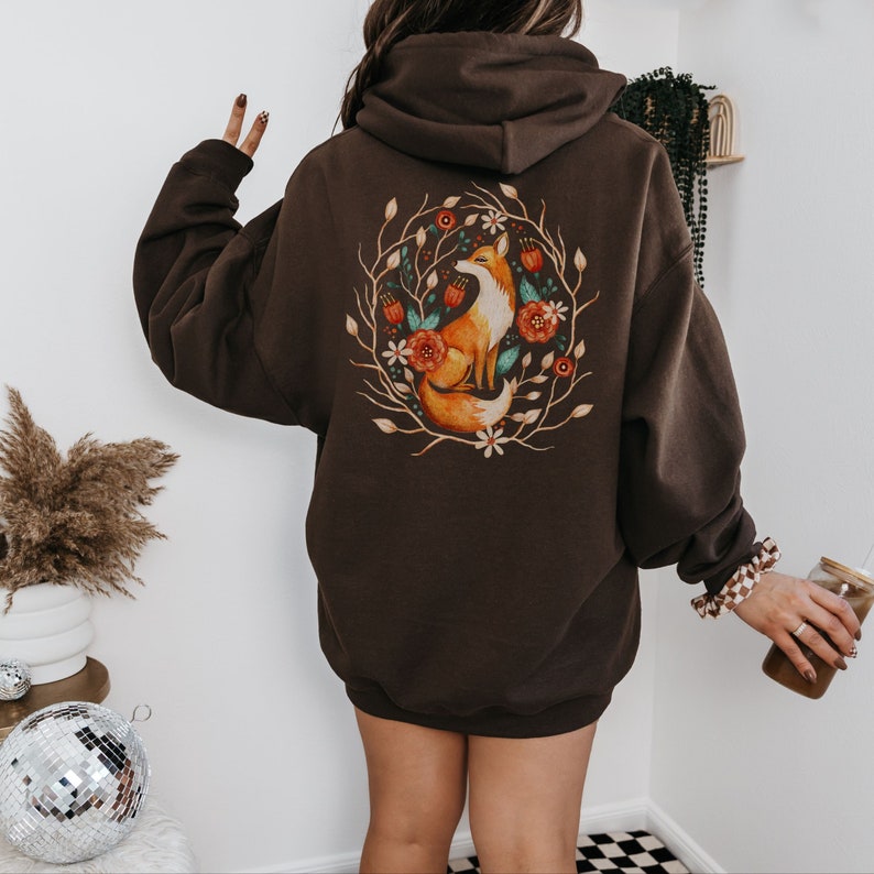 May include: A brown hooded sweatshirt with a graphic of a fox sitting in a circle of flowers. The fox is orange and white with a bushy tail. The flowers are red, orange, and yellow.