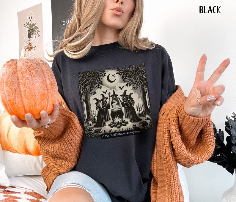 Puede incluir: Una camiseta negra con un gr&aacute;fico de tres brujas de pie alrededor de un caldero en un bosque. El texto "Sisters of magic & mayhem" est&aacute; impreso debajo del gr&aacute;fico.