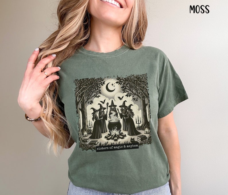 Puede incluir: Una camiseta verde con un estampado gr&aacute;fico de tres brujas de pie alrededor de un caldero en un entorno de bosque. El texto "sisters of magic & mayhem" est&aacute; impreso debajo del gr&aacute;fico.