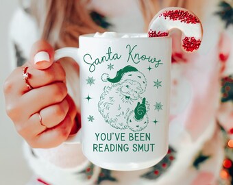 May include: White ceramic mug with the text "Santa Knows You've Been Reading Smut" and a green Santa Claus illustration. A candy cane cookie with red sprinkles rests on the rim. The mug is filled with a white substance.