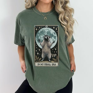 May include: A green t-shirt with a graphic of a raccoon standing on its hind legs with its paws raised in the air. The raccoon is in front of a full moon and stars. The text "IN MY FERAL ERA" is printed below the graphic.