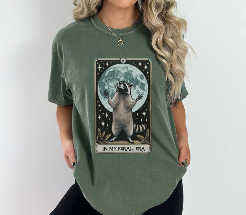 May include: A green t-shirt with a graphic of a raccoon standing on its hind legs with its paws raised in the air. The raccoon is in front of a full moon and stars. The text "IN MY FERAL ERA" is printed below the graphic.