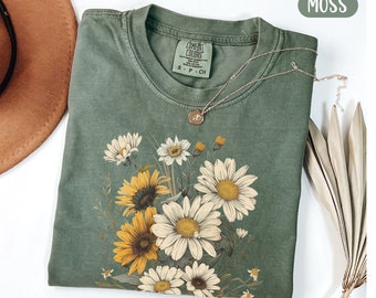 May include: A green t-shirt with a floral print featuring sunflowers and daisies. The shirt has a distressed look and the text "COMFORT COLORS" is visible on the tag.