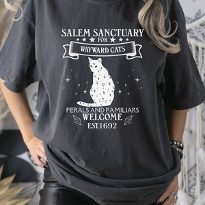 Może przedstawiać: Czarny T-shirt z białym nadrukiem graficznym, na kt&oacute;rym widnieje napis "Salem Sanctuary for Wayward Cats" z ilustracją kota i tekstem "Ferals and Familiars Welcome Est. 1692".