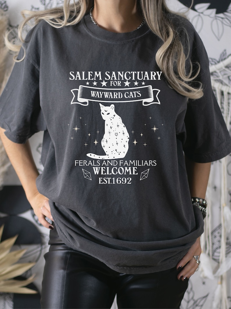 Może przedstawiać: Czarny T-shirt z białym nadrukiem graficznym, na kt&oacute;rym widnieje napis "Salem Sanctuary for Wayward Cats" z ilustracją kota i tekstem "Ferals and Familiars Welcome Est. 1692".