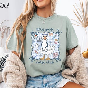 May include: A light green t-shirt featuring a cartoon goose wearing a nurse's cap and stethoscope, holding a drink. The design includes the text "silly goose nurse club" within a blue and white gingham frame, with medical-themed illustrations.