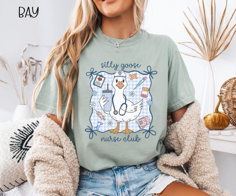May include: A light green t-shirt featuring a cartoon goose wearing a nurse's cap and stethoscope, holding a drink. The design includes the text "silly goose nurse club" within a blue and white gingham frame, with medical-themed illustrations.