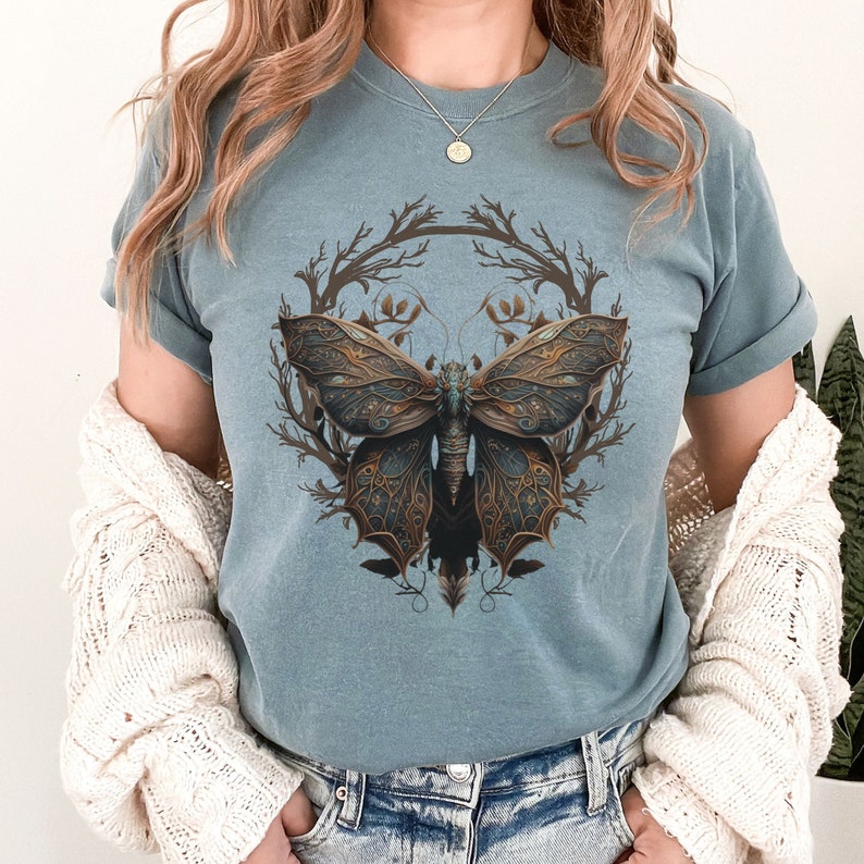 May include: A light blue t-shirt with a graphic of a detailed, intricate butterfly with brown and gold accents. The butterfly is surrounded by a stylized branch design.