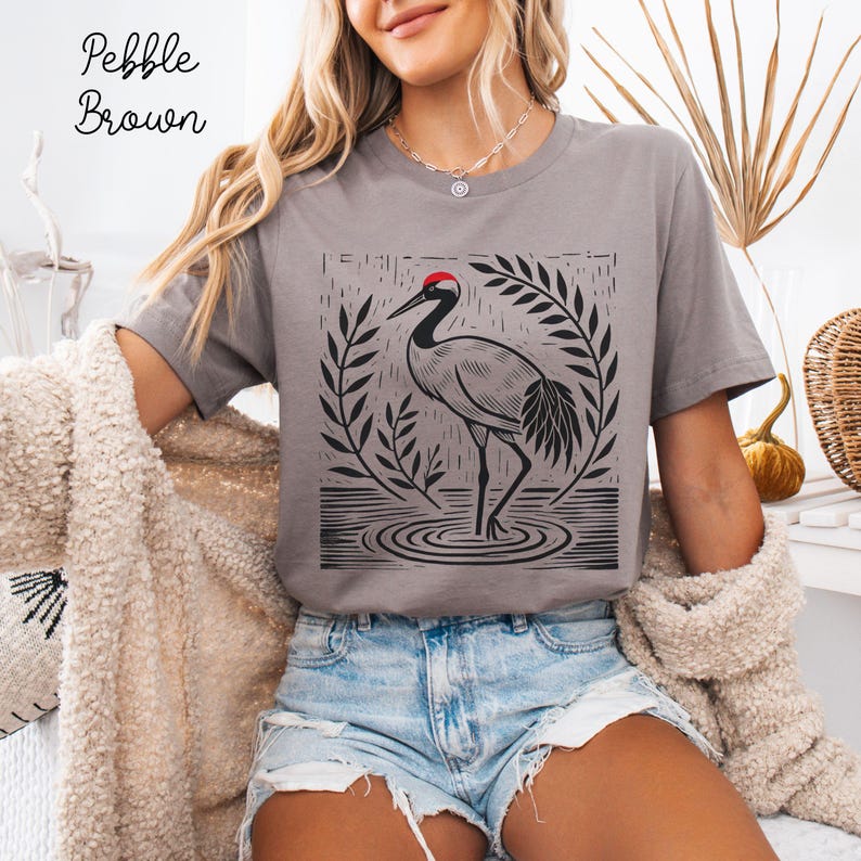 May include: A pebble brown t-shirt featuring a black graphic print of a crane standing in water, surrounded by a leaf-like border. The crane has a distinct red spot on its head. The shirt has short sleeves and a crew neckline, with the text Pebble Brown.