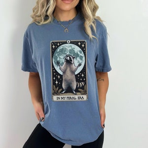 May include: A blue t-shirt with a graphic of a raccoon standing on its hind legs with its paws raised in the air. The raccoon is in front of a full moon and stars. The text "IN MY FERAL ERA" is printed below the raccoon.