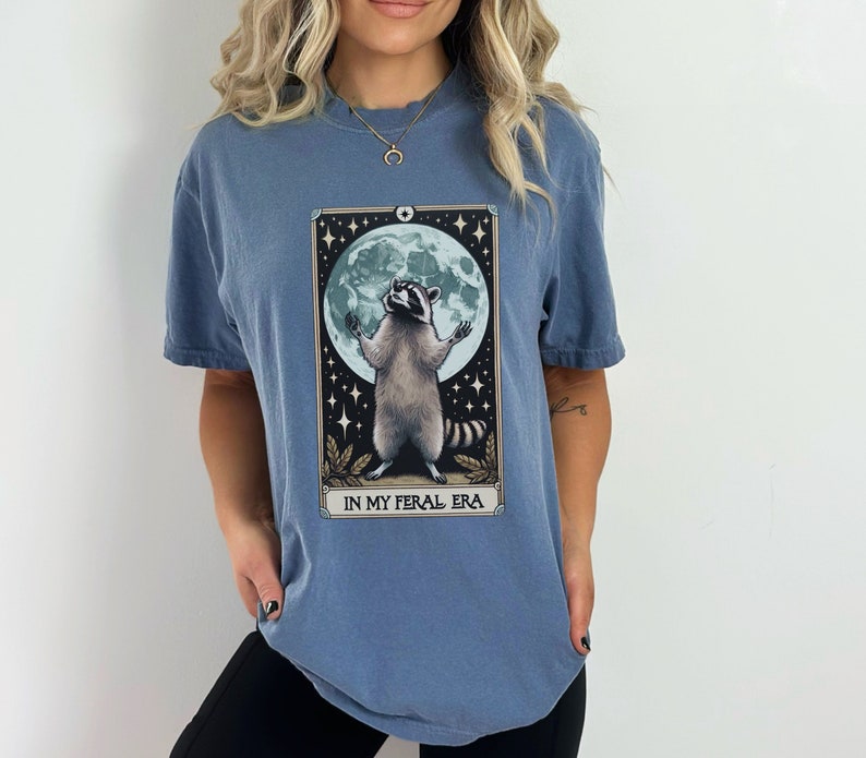 May include: A blue t-shirt with a graphic of a raccoon standing on its hind legs with its paws raised in the air. The raccoon is in front of a full moon and stars. The text "IN MY FERAL ERA" is printed below the raccoon.