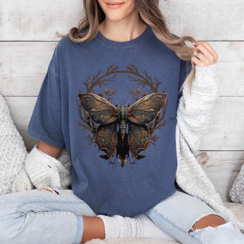 May include: A navy blue t-shirt with a graphic of a detailed, intricate butterfly with gold and green accents. The butterfly is surrounded by a wreath of branches.