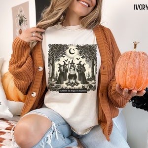 Puede incluir: Una camiseta blanca con una ilustraci&oacute;n vintage de tres brujas en un entorno de bosque. Las brujas est&aacute;n de pie alrededor de un caldero y hay un gato negro en primer plano. El texto "Sisters of magic & mayhem" est&aacute; impreso debajo de la ilustraci&oacute;n.