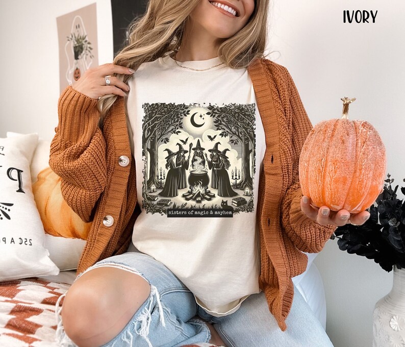 Puede incluir: Una camiseta blanca con una ilustraci&oacute;n vintage de tres brujas en un entorno de bosque. Las brujas est&aacute;n de pie alrededor de un caldero y hay un gato negro en primer plano. El texto "Sisters of magic & mayhem" est&aacute; impreso debajo de la ilustraci&oacute;n.