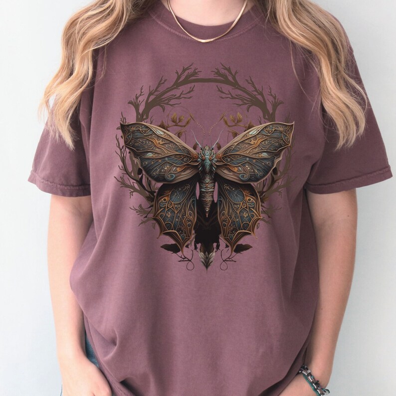 May include: A mauve colored t-shirt with a graphic print of a detailed, intricate butterfly with gold and blue accents. The butterfly is surrounded by a circular design of brown branches.