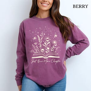 May include: Berry-coloured long-sleeve shirt with a floral and celestial design. The graphic features an open book with flowers and the text "Just One More Chapter" in a script font. The shirt is made of a soft material.