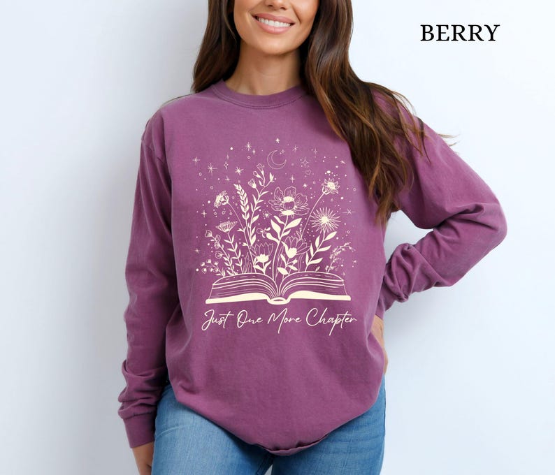 May include: Berry-coloured long-sleeve shirt with a floral and celestial design. The graphic features an open book with flowers and the text "Just One More Chapter" in a script font. The shirt is made of a soft material.