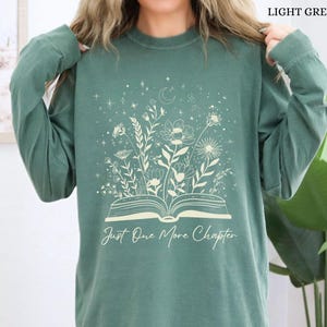 May include: Light green long-sleeved shirt with a floral and celestial design. The design features an open book with flowers and stars, and the text "Just One More Chapter". The shirt is a solid light green colour.