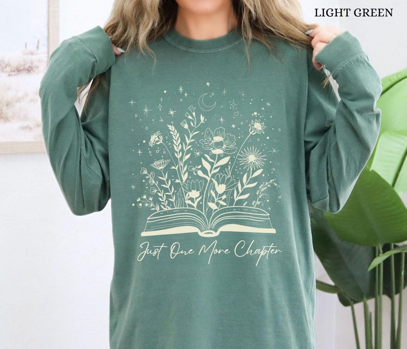 May include: Light green long-sleeved shirt with a floral and celestial design. The design features an open book with flowers and stars, and the text "Just One More Chapter". The shirt is a solid light green colour.