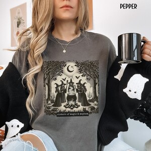 Puede incluir: Una sudadera gris con un estampado gr&aacute;fico de tres brujas de pie alrededor de un caldero en un bosque. El texto "sisters of magic & mayhem" est&aacute; impreso debajo del gr&aacute;fico.
