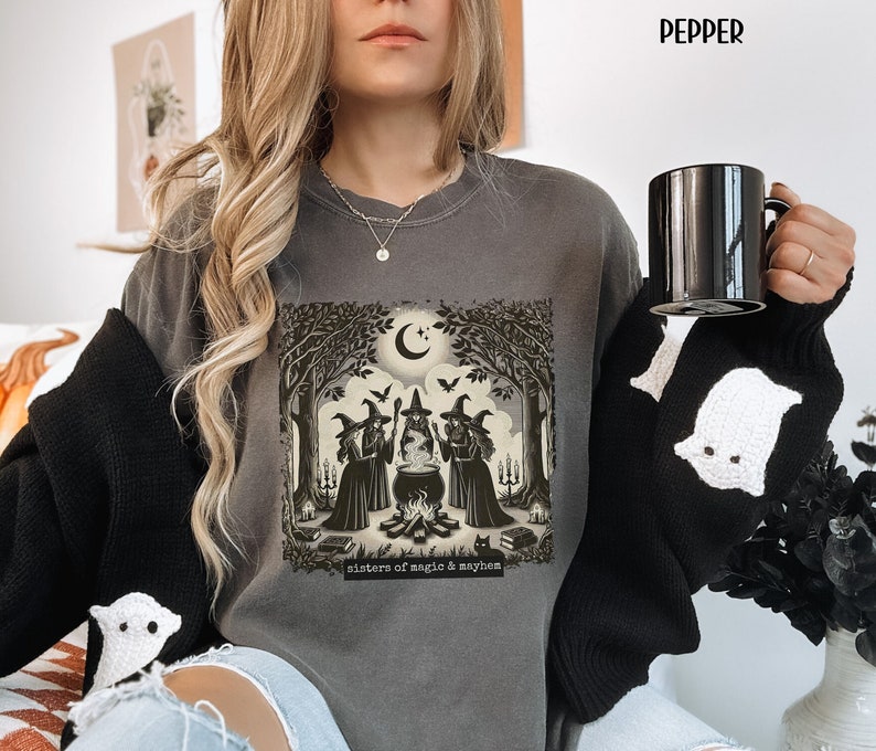 Puede incluir: Una sudadera gris con un estampado gr&aacute;fico de tres brujas de pie alrededor de un caldero en un bosque. El texto "sisters of magic & mayhem" est&aacute; impreso debajo del gr&aacute;fico.