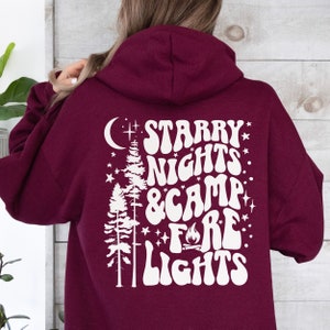 May include: A maroon hoodie with a white graphic design on the back. The graphic features a crescent moon, two pine trees, stars, and the text "Starry Nights & Camp Fire Lights" in a retro font.