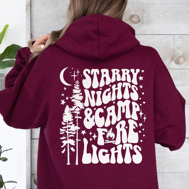 May include: A maroon hoodie with a white graphic design on the back. The graphic features a crescent moon, two pine trees, stars, and the text "Starry Nights & Camp Fire Lights" in a retro font.
