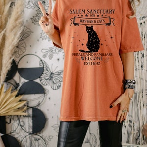 Może przedstawiać: Pomarańczowy t-shirt z czarnym nadrukiem graficznym kota z gwiazdami. Tekst na koszulce brzmi "Salem Sanctuary for Wayward Cats" i "Ferals and Familiars Welcome Est. 1692".