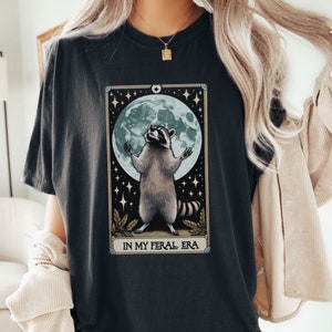 May include: A black t-shirt with a graphic of a raccoon standing on its hind legs with its paws raised in the air. The raccoon is in front of a full moon with stars in the background. The text "In My Feral Era" is printed below the raccoon.