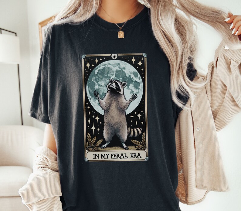 May include: A black t-shirt with a graphic of a raccoon standing on its hind legs with its paws raised in the air. The raccoon is in front of a full moon with stars in the background. The text "In My Feral Era" is printed below the raccoon.