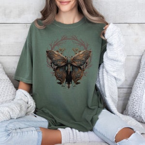 May include: A green t-shirt with a graphic of a detailed, intricate, steampunk-style butterfly with gold and brown accents. The butterfly is surrounded by a wreath of branches and leaves.