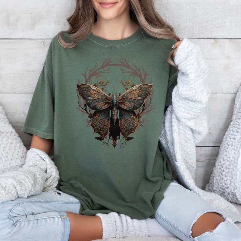 May include: A green t-shirt with a graphic of a detailed, intricate, steampunk-style butterfly with gold and brown accents. The butterfly is surrounded by a wreath of branches and leaves.