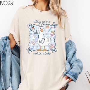 May include: Ivory-colored t-shirt featuring a cartoon goose wearing a nurse's hat and stethoscope, holding a drink and medical supplies. The design includes the text "silly goose nurse club" within a blue gingham frame.
