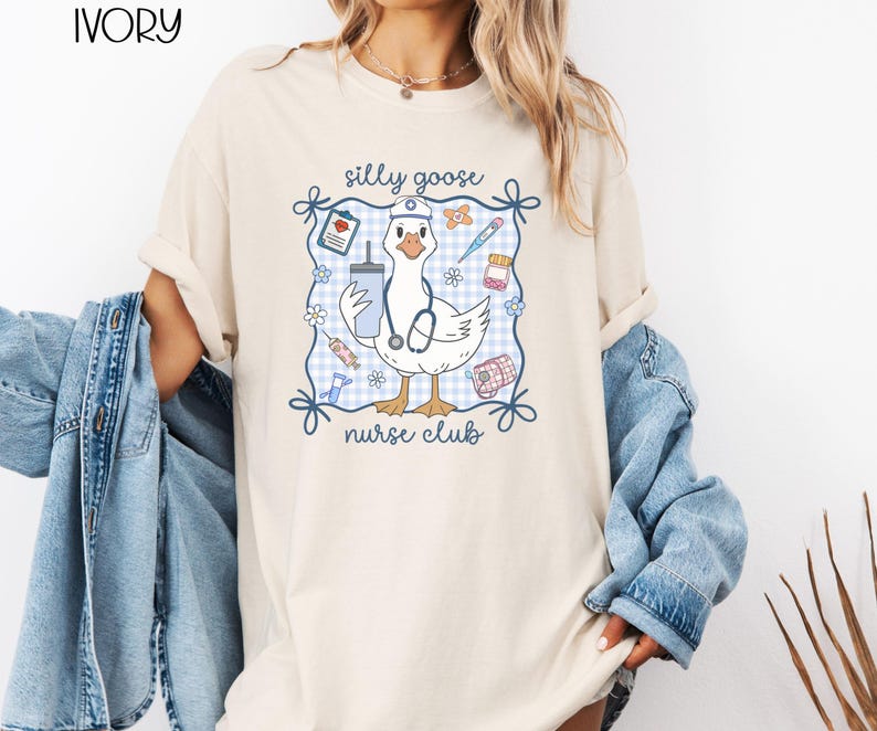 May include: Ivory-colored t-shirt featuring a cartoon goose wearing a nurse's hat and stethoscope, holding a drink and medical supplies. The design includes the text "silly goose nurse club" within a blue gingham frame.