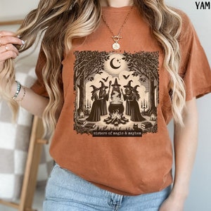 Puede incluir: Una camiseta marr&oacute;n con un estampado gr&aacute;fico de tres brujas en un entorno de bosque. Las brujas est&aacute;n de pie alrededor de un caldero y hay un gato negro en primer plano. El texto "sisters of magic & mayhem" est&aacute; impreso debajo del gr&aacute;fico.