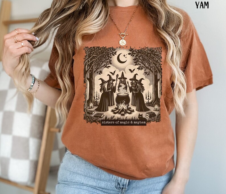 Puede incluir: Una camiseta marr&oacute;n con un estampado gr&aacute;fico de tres brujas en un entorno de bosque. Las brujas est&aacute;n de pie alrededor de un caldero y hay un gato negro en primer plano. El texto "sisters of magic & mayhem" est&aacute; impreso debajo del gr&aacute;fico.